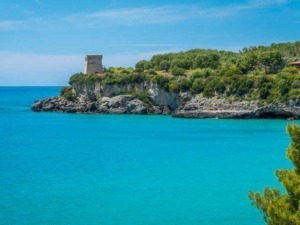 Spiaggia con acque cristalline, scogliera verdeggiante e antica torre di avvistamento, perfetta destinazione per relax e turismo in Italia. Panorama mozzafiato soleggiato e nature incontaminata.