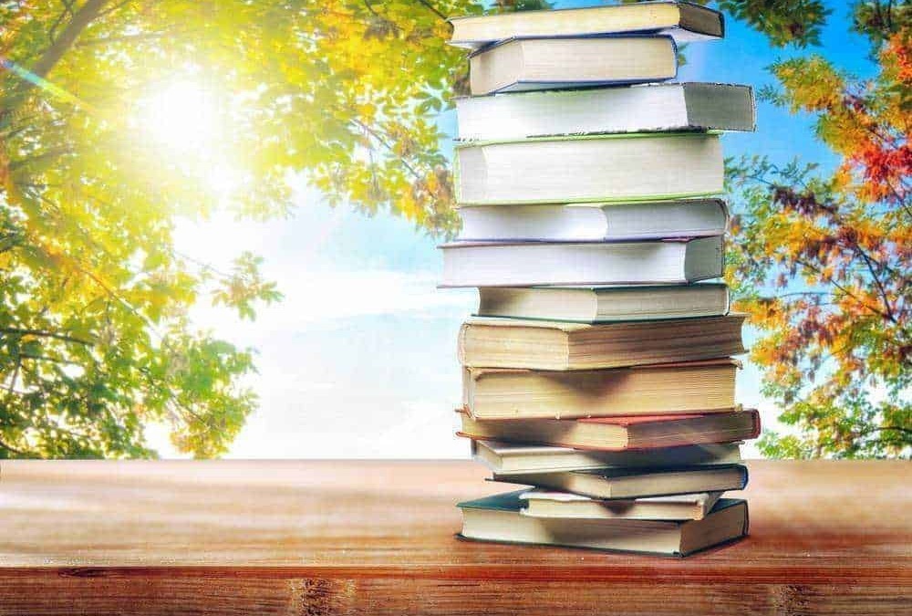Pila di libri su un tavolo di legno con sfondo di alberi in autunno e cielo soleggiato.
