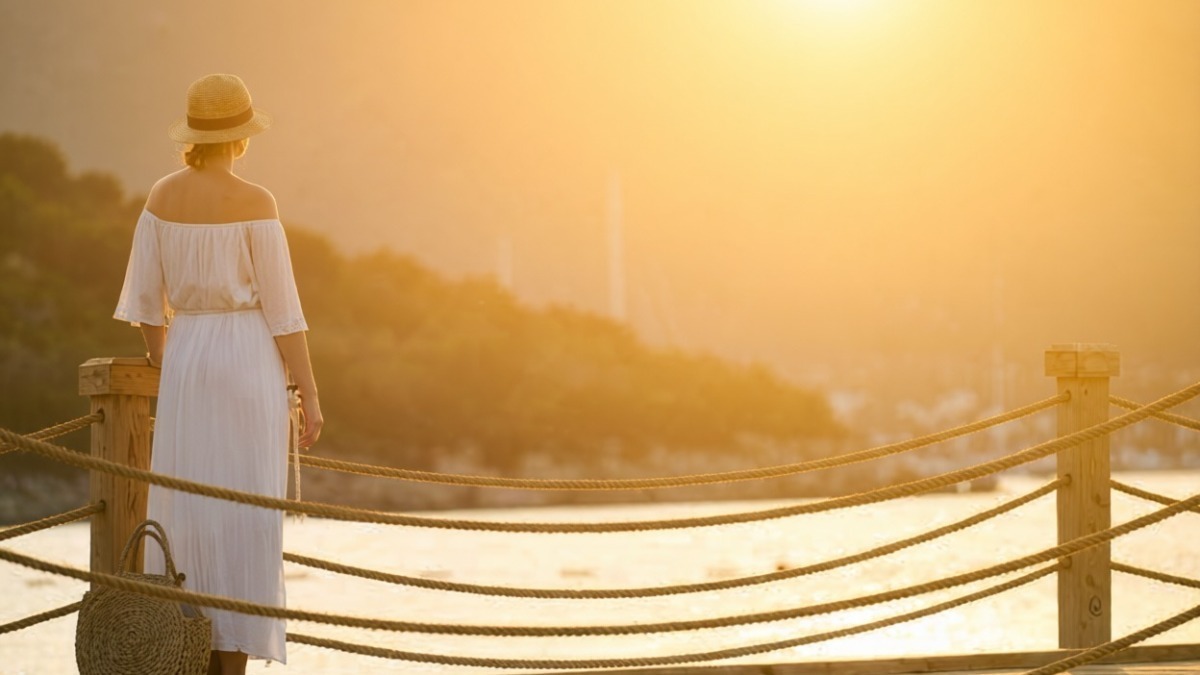 Vecchia donna con cappello e vestito bianco che guarda il tramonto dal pontile sul fiume in un paesaggio naturale, atmosfera serena e rilassante.