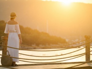 Vecchia donna con cappello e vestito bianco che guarda il tramonto dal pontile sul fiume in un paesaggio naturale, atmosfera serena e rilassante.