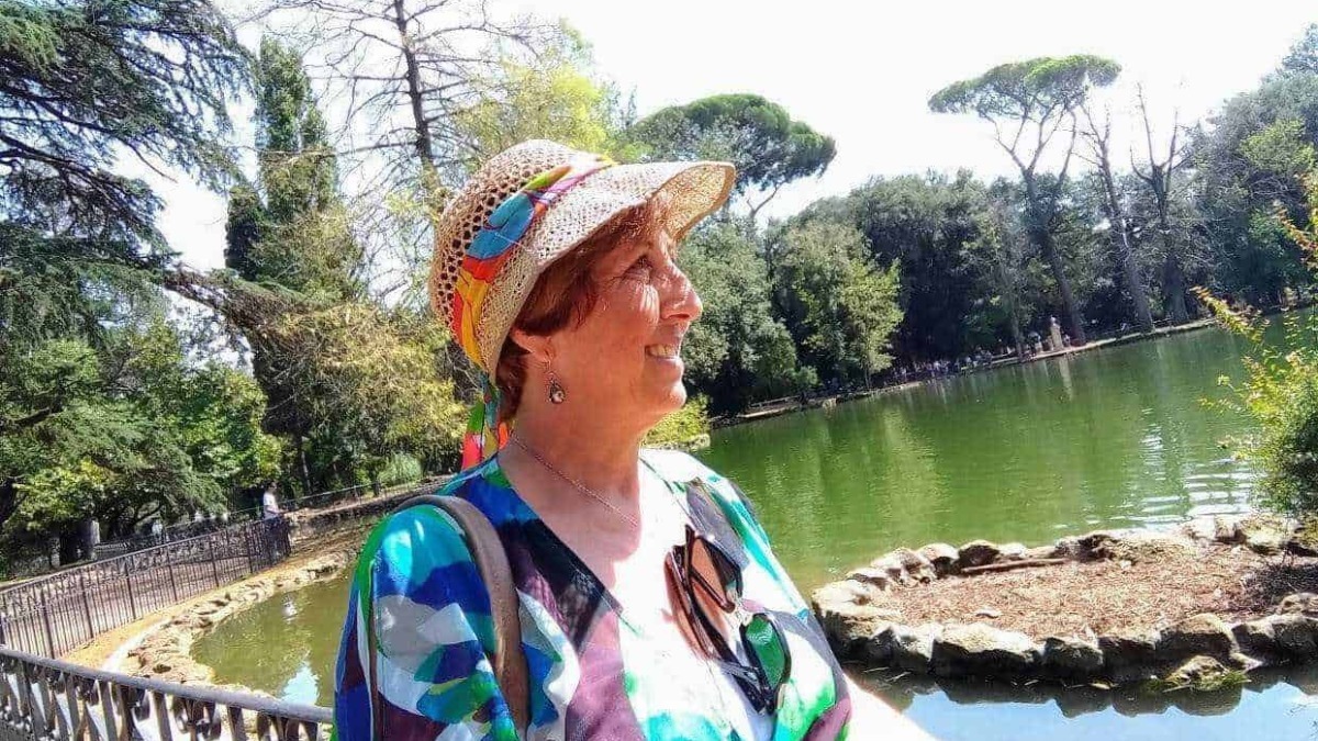 Femminile anziana sorridente con cappello di paglia colorato che si gode una passeggiata al parco vicino a uno stagno in una giornata soleggiata.
