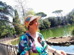 Femminile anziana sorridente con cappello di paglia colorato che si gode una passeggiata al parco vicino a uno stagno in una giornata soleggiata.