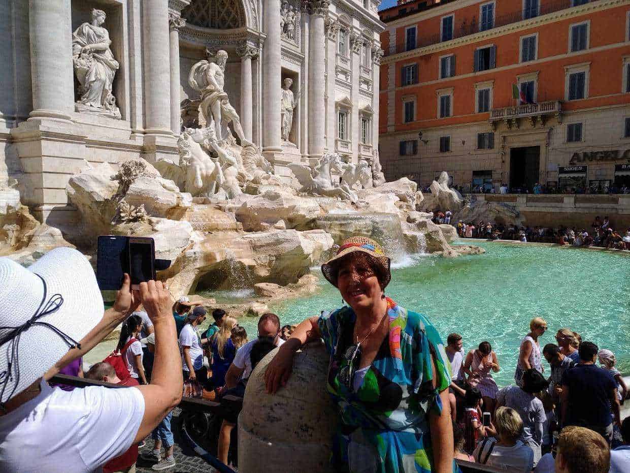 Femminile turista sorridente davanti alla Fontana di Trevi a Roma, Italia, mentre scatta una foto, con molti visitatori e architetture storiche sullo sfondo.
