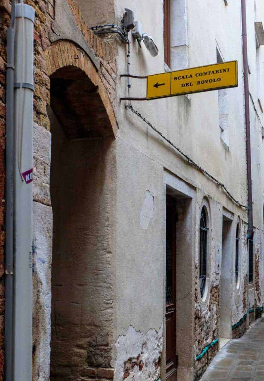 Vecchio vicolo medievale con segnale stradale giallo "Scala Contarini del Bovalo" e facciata in pietra e intonaco decorativo.