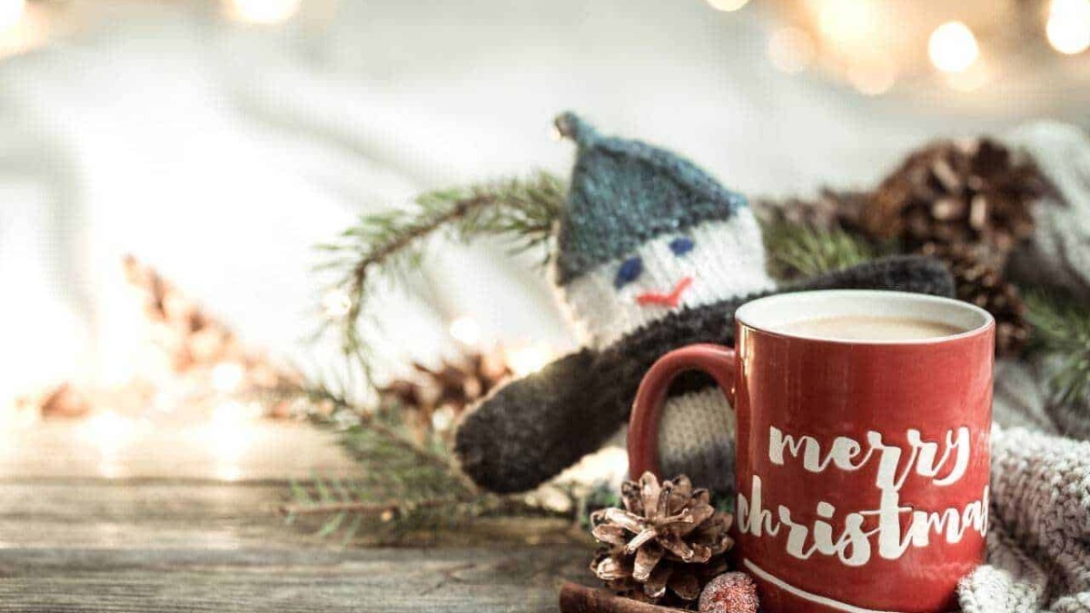Tazza di cioccolata calda con dedica natalizia, decorata con elementi di Natale come pigne, corteccia di canna e pupazzo di neve di lana, atmosfera festiva e calda per le festività natalizie.