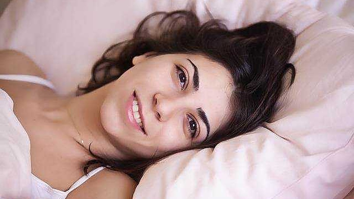 Sonno poco profondo: Bellissima donna con capelli scuri e sorriso naturale, che riposa su un letto con cuscini rosa, in un ambiente luminoso e accogliente.