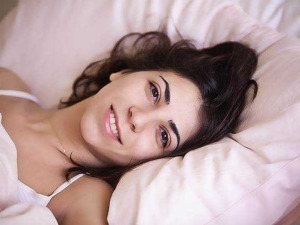 Sonno poco profondo: Bellissima donna con capelli scuri e sorriso naturale, che riposa su un letto con cuscini rosa, in un ambiente luminoso e accogliente.