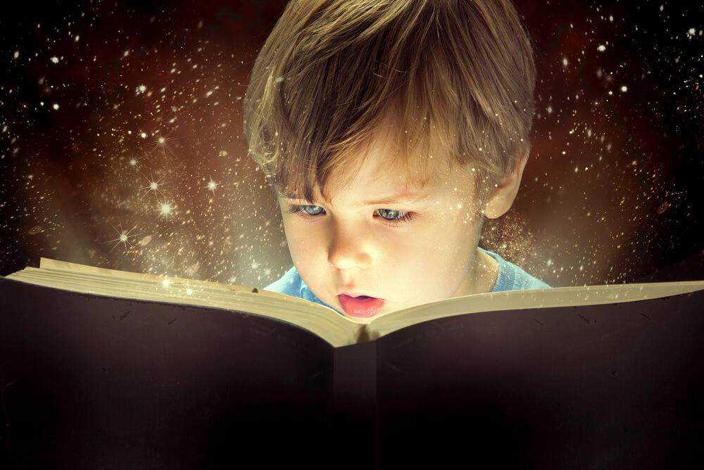 Immagine di un giovane bambino con capelli castani chiari che legge un libro magico circondato da stelle scintillanti e particelle luminose, creando un'atmosfera fantastica di scoperta e meraviglia.