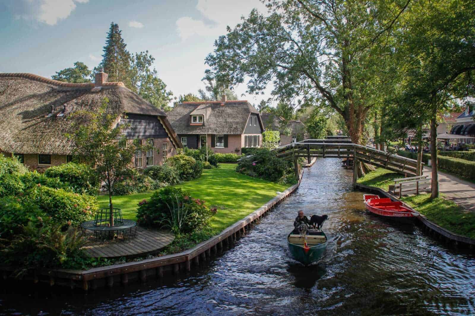 13 Città con canali nel mondo da vedere 4 Casa olandese in stile tradizionale lungo un canale con giardino curato e barche in acqua, in un paesaggio pittoresco tipico delle città olandesi, con alberi verdi e cielo sereno.
