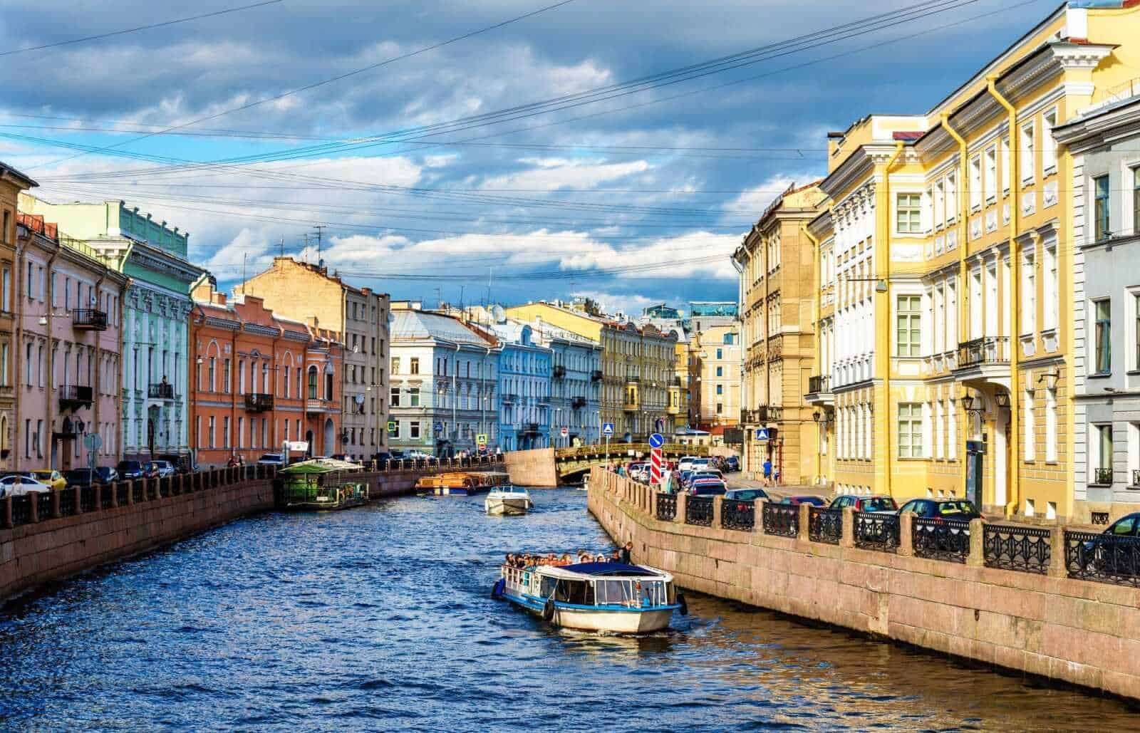 13 Città con canali nel mondo da vedere 10 Colorate case storiche lungo il canale di San Pietroburgo, Russia, con barche che navigano sull'acqua sotto un cielo nuvoloso. Edifici colorati e atmosfera vivace in centro città.