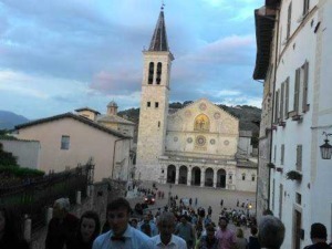 Campanile e facciata della chiesa di San Michele a Borgo San Michele, Italia, con molte persone e un cielo nuvoloso sullo sfondo.