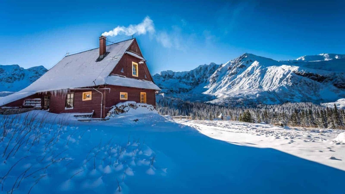 Casetta di legno rossa coperta di neve con montagne innevate sullo sfondo in un paesaggio invernale; luogo ideale per appunti sul blog di viaggio.