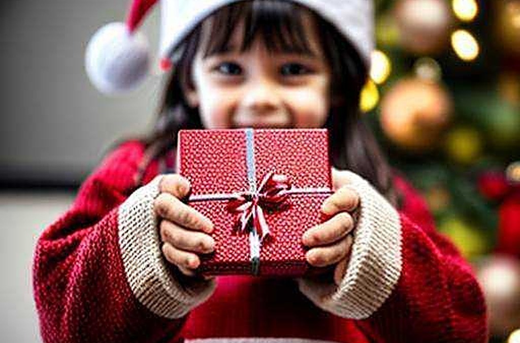 Regali di Natale per bambini con cappello di Babbo Natale, sorriso e scatola regalo rossa, atmosfera festiva e decorazioni natalizie alle spalle.