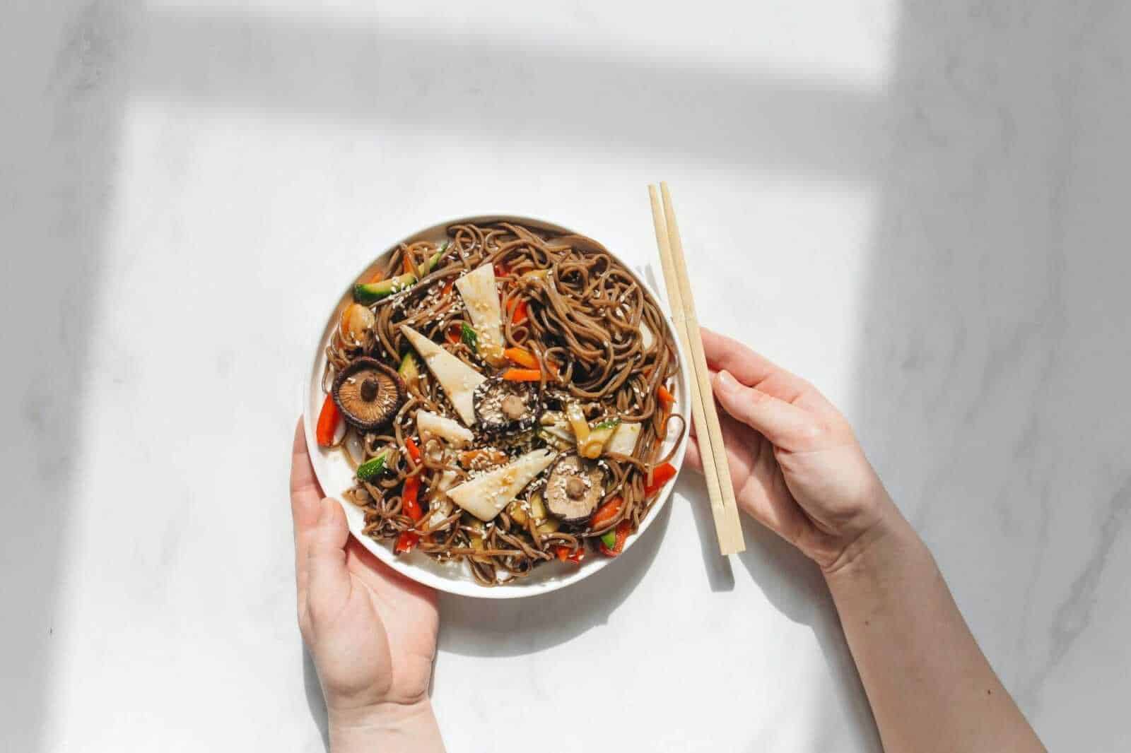 Spaghetti di soba con verdure e funghi, piatto giapponese di noodles integrali, servito in una ciotola bianca con bacchette, stile minimalista, presentato su sfondo bianco.