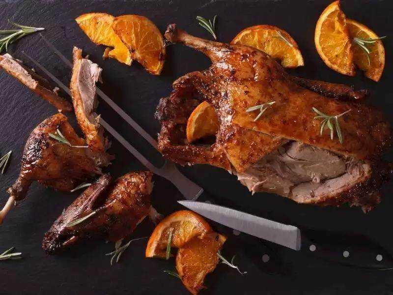 Succulent roasted lamb with orange slices and fresh herbs on black slate background.