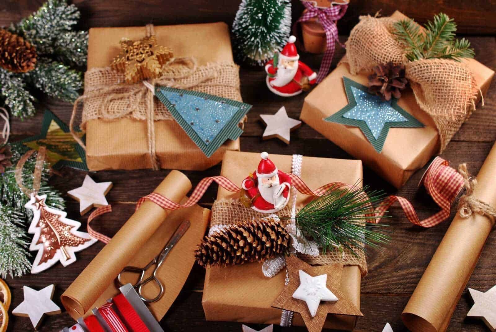 Pacchi regalo di Natale avvolti in carta kraft con decorazioni natalizie, fiocco di neve, rametti di pino, e figure di Babbo Natale, su tavolo di legno per preparazioni festive.