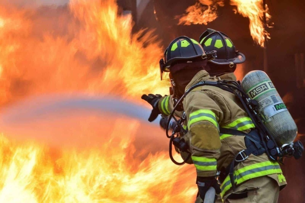 Sensan firefighting firefighter wearing protective gear and helmet extinguishing a blaze with a fire hose during a wildfire emergency.