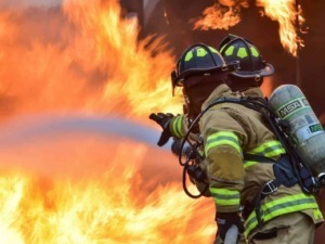 Sensan firefighting firefighter wearing protective gear and helmet extinguishing a blaze with a fire hose during a wildfire emergency.