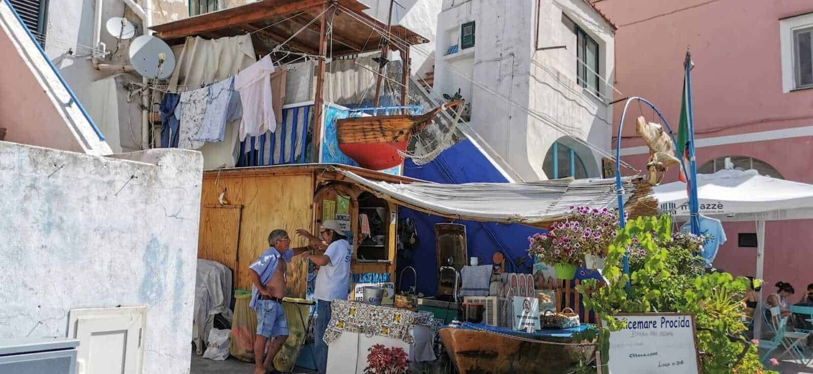 Vicolo colorato con bancarella di street food e persone che conversano, case pittoresche con facciate in tonalità pastello, panni stesi, e decorazioni floreali in un quartiere caratteristico di pescatori.