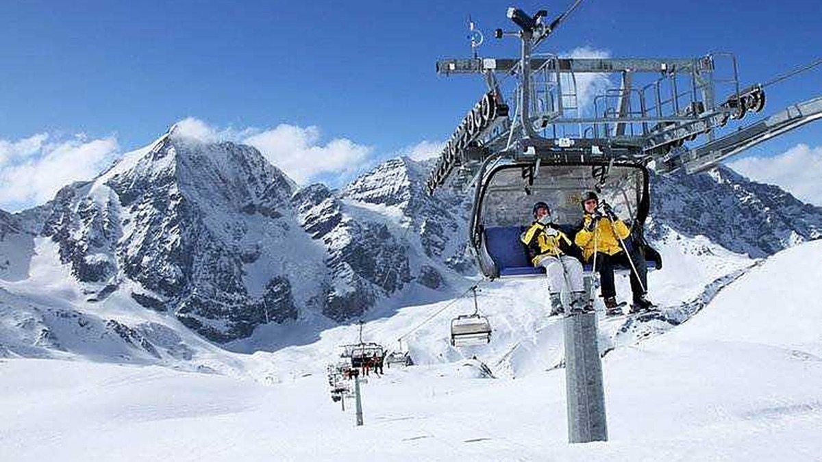 Sciatori sulle piste innevate delle Alpi durante una bella giornata di sole, con impianto di ski lift in vista e montagne imponenti sullo sfondo.