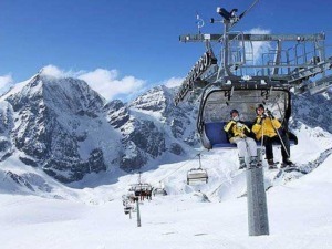 Sciatori sulle piste innevate delle Alpi durante una bella giornata di sole, con impianto di ski lift in vista e montagne imponenti sullo sfondo.