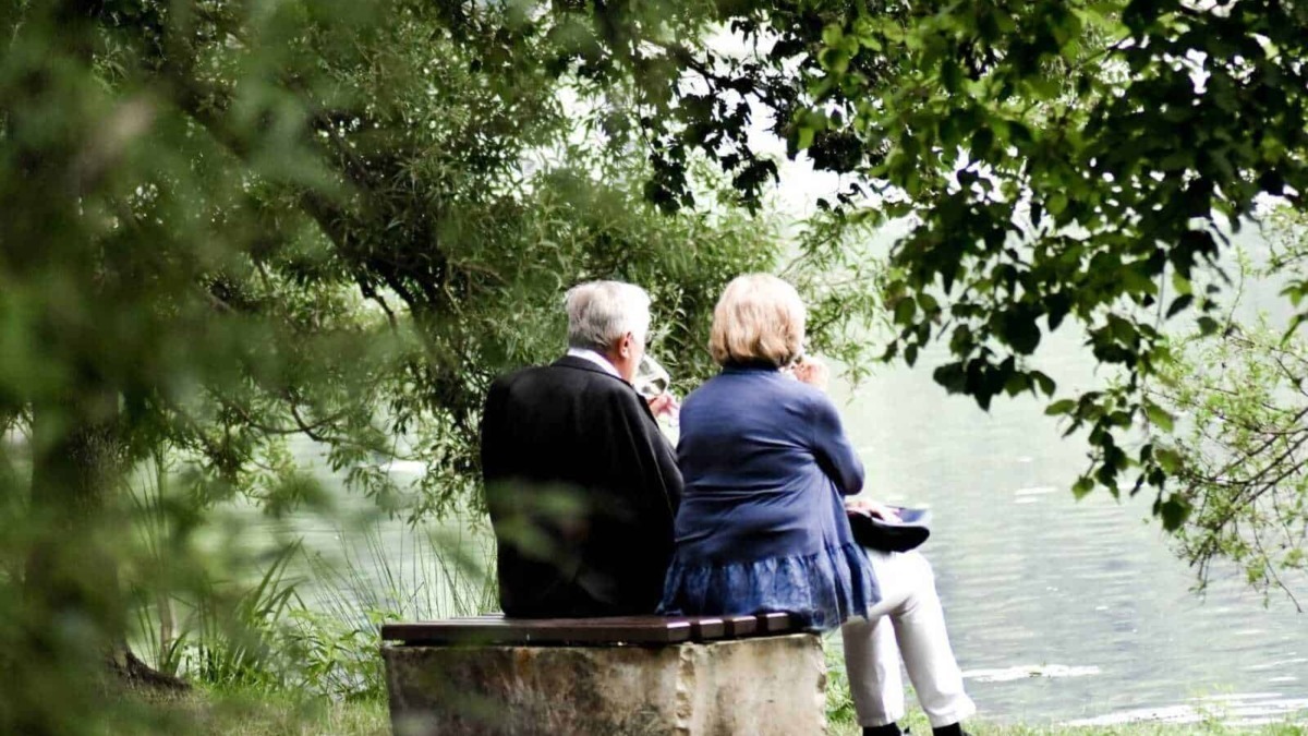 Relaxing elderly couple sitting on a wooden bench by the lake, surrounded by lush green foliage, enjoying a peaceful moment in nature.
