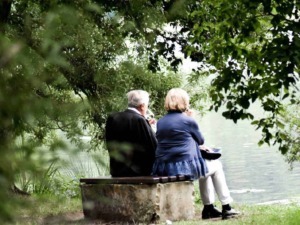 Relaxing elderly couple sitting on a wooden bench by the lake, surrounded by lush green foliage, enjoying a peaceful moment in nature.