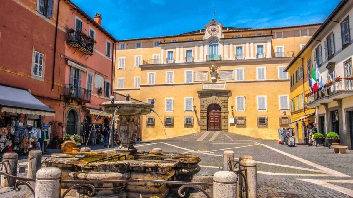 Fontana storica in una piazza di un centro storico italiano con edifici colorati sullo sfondo sotto un cielo blu.