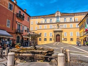 Fontana storica in una piazza di un centro storico italiano con edifici colorati sullo sfondo sotto un cielo blu.