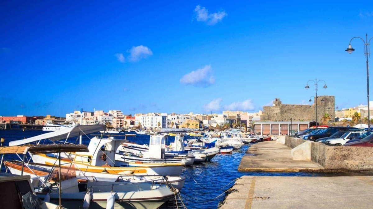 Immagine di un porticciolo con barche a vela e motore, un'area di parcheggio e un castello storico sullo sfondo, in una vivace città costiera italiana.