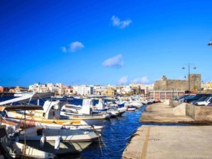 Immagine di un porticciolo con barche a vela e motore, un'area di parcheggio e un castello storico sullo sfondo, in una vivace città costiera italiana.
