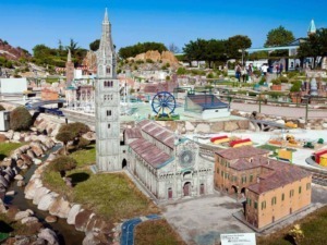 Miniatura di un parco con modelli in miniatura di edifici e attrazioni, tra cui una chiesa e una torre, visitato da persone, con dettagli realistici e ambientazione all'aperto sotto un cielo azzurro.