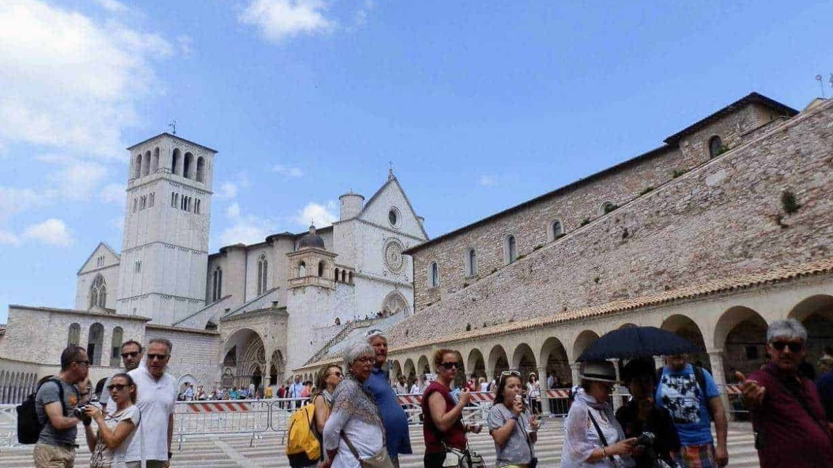Fila di turisti davanti al Duomo di San Francesco in Assisi, Italia, con un cielo azzurro e nuvole sparse, visitando uno dei luoghi storici più importanti della regione Umbria.