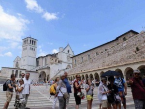 Fila di turisti davanti al Duomo di San Francesco in Assisi, Italia, con un cielo azzurro e nuvole sparse, visitando uno dei luoghi storici più importanti della regione Umbria.