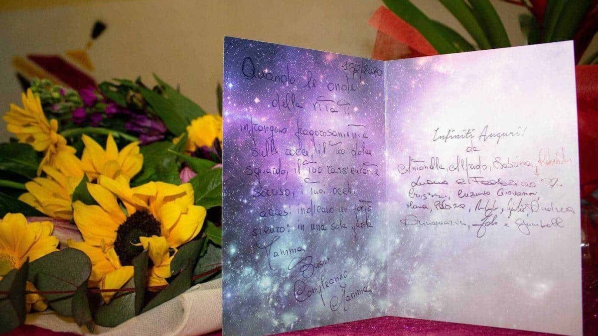 Fiocco di biglietto con sfondo stellato tra bouquet di fiori gialli e viola, messaggio di auguri e dediche sentimentali, decorazioni floreali, atmosfera calda e poetica.