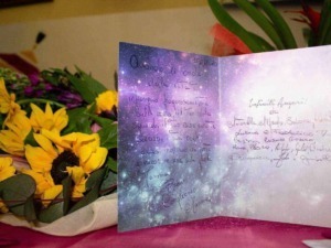 Fiocco di biglietto con sfondo stellato tra bouquet di fiori gialli e viola, messaggio di auguri e dediche sentimentali, decorazioni floreali, atmosfera calda e poetica.