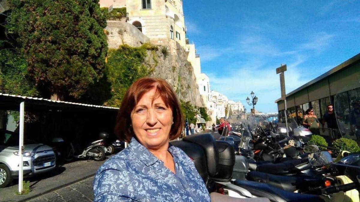 Vita quotidiana a Positano, Italia, con una donna sorridente di fronte a un lungomare affollato di scooter, con case colorate e il mare sullo sfondo, ambiente vivace e turistico.