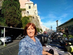 Vita quotidiana a Positano, Italia, con una donna sorridente di fronte a un lungomare affollato di scooter, con case colorate e il mare sullo sfondo, ambiente vivace e turistico.