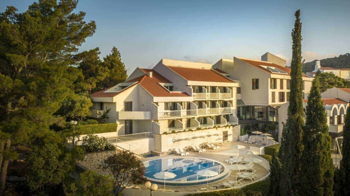 Elegante hotel con piscina all'aperto circondata da alberi e vegetazione, edifici moderni con tetti in terracotta, ideale per vacanze rilassanti in una località turistica italiana.