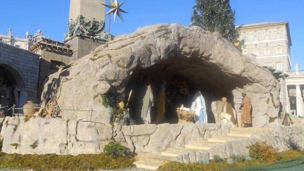 Croce di Natale con presepe vivente sotto una grotta di pietra, decorata con un albero di Natale e figure religiose, situata in un contesto urbano storico.