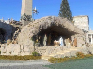 Croce di Natale con presepe vivente sotto una grotta di pietra, decorata con un albero di Natale e figure religiose, situata in un contesto urbano storico.