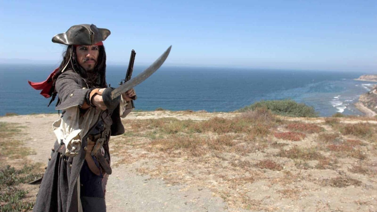 Corsaro dei Caraibi con spada e pistola, vista dell'oceano e scogliera sullo sfondo, costume da pirata autentico, avventura e divertimento piratesco, ambientazione di mare aperto e natura selvaggia.