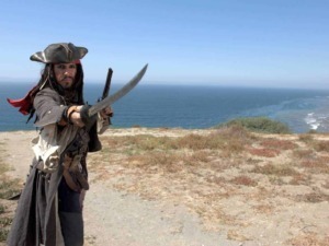 Corsaro dei Caraibi con spada e pistola, vista dell'oceano e scogliera sullo sfondo, costume da pirata autentico, avventura e divertimento piratesco, ambientazione di mare aperto e natura selvaggia.