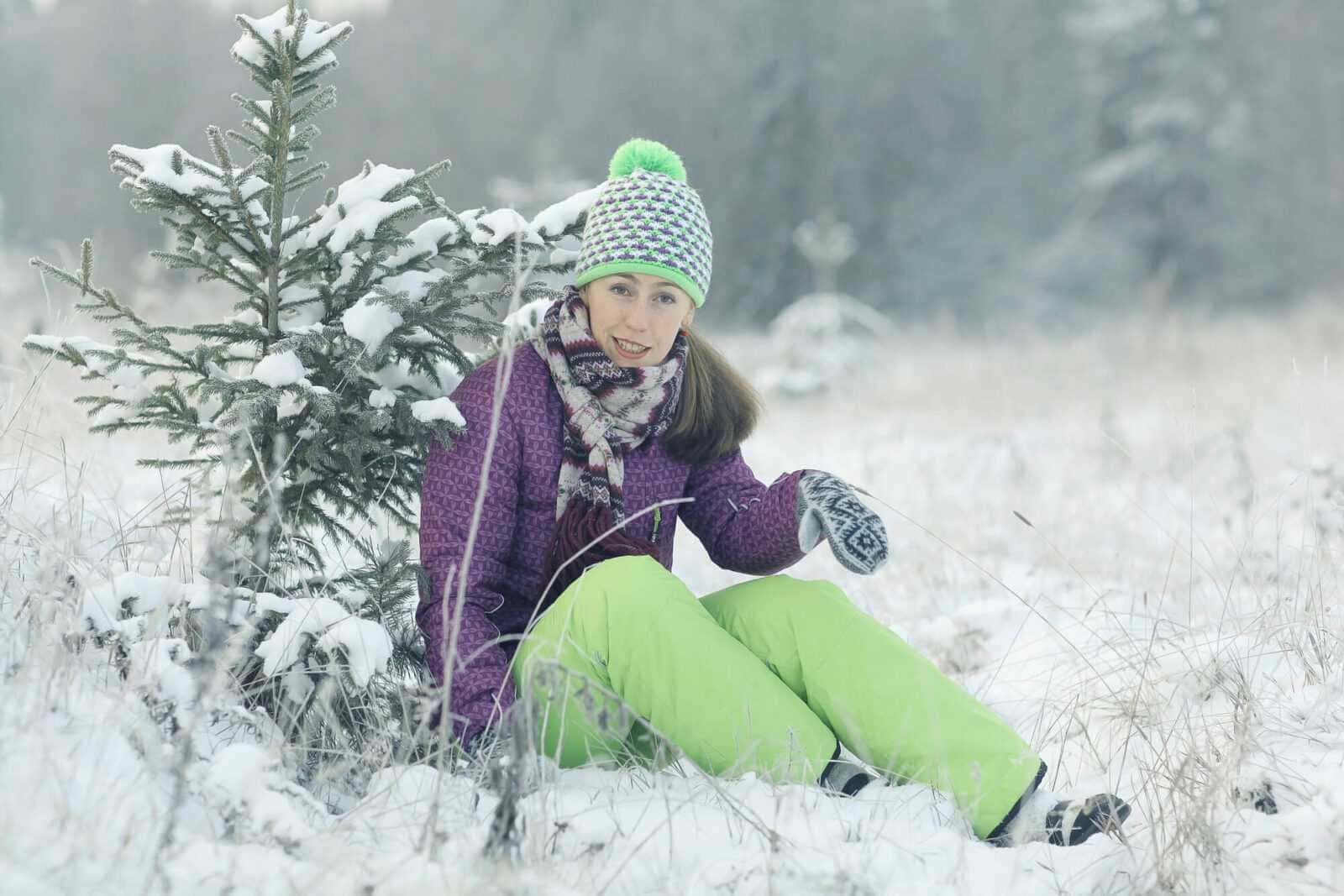 Nevicata invernale: ragazza sorridente con abbigliamento caldo e muscolare seduta nel paesaggio innevato, circondata da alberi di pino e natura, ideale per escursioni in montagna e attività outdoor in inverno.