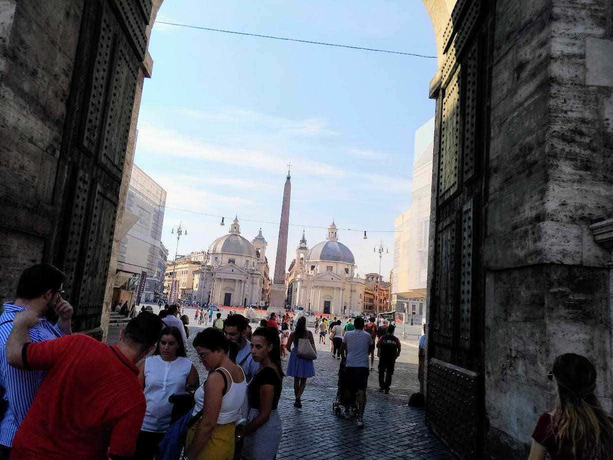 Passeggiata nel cuore di Roma con vista su Piazza del Popolo, le chiese gemelle e l'obelisco nel centro, tra turisti e cittadini che ammirano l'arte e la storia della città eterna.