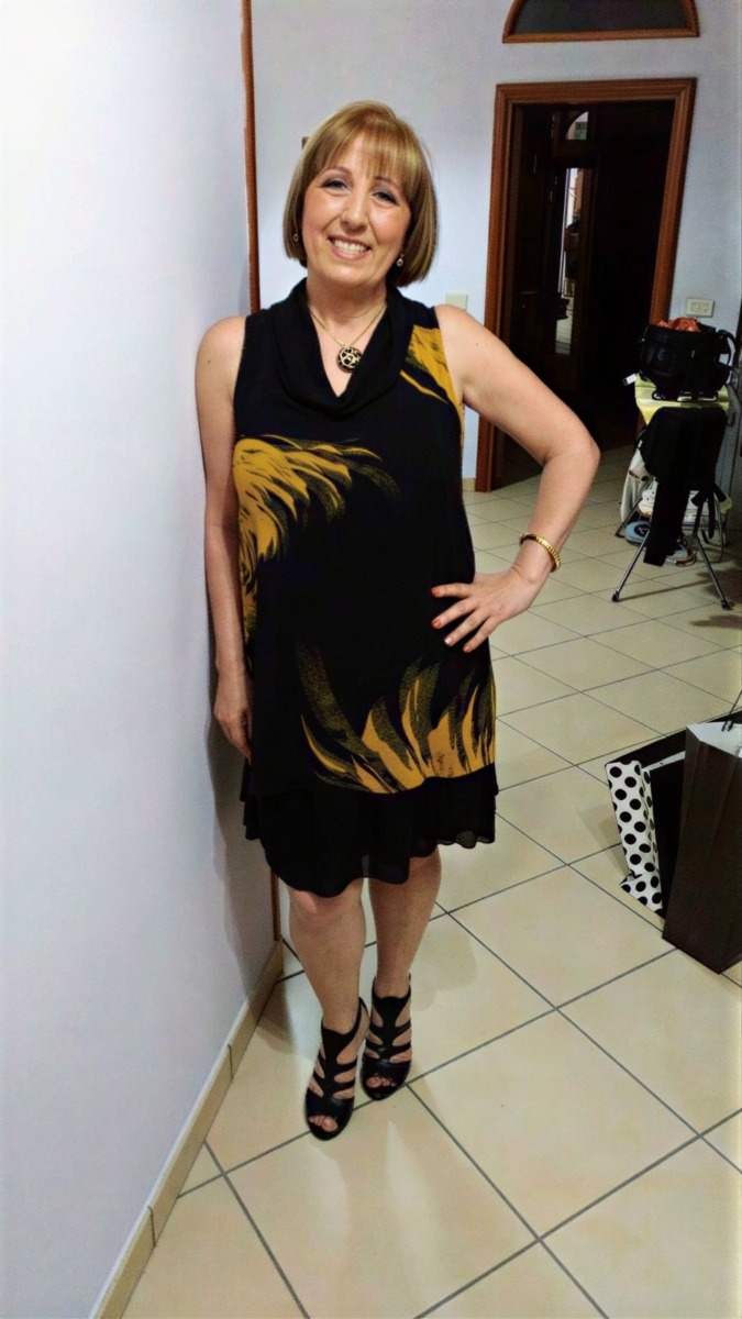 Sanremo 2012 dietro le quinte: Elegante donna in abito nero e giallo con scarpe col tacco, sorridente e in posa all’interno di un ambiente domestico, perfetta per immagini di moda e stile di vita.