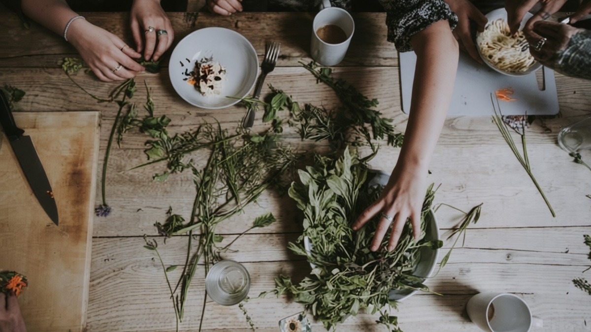 Un gruppo di persone che prepara un'insalata con verdure fresche su un tavolo di legno ricco di ingredienti e utensili da cucina, ambiente conviviale e informale.