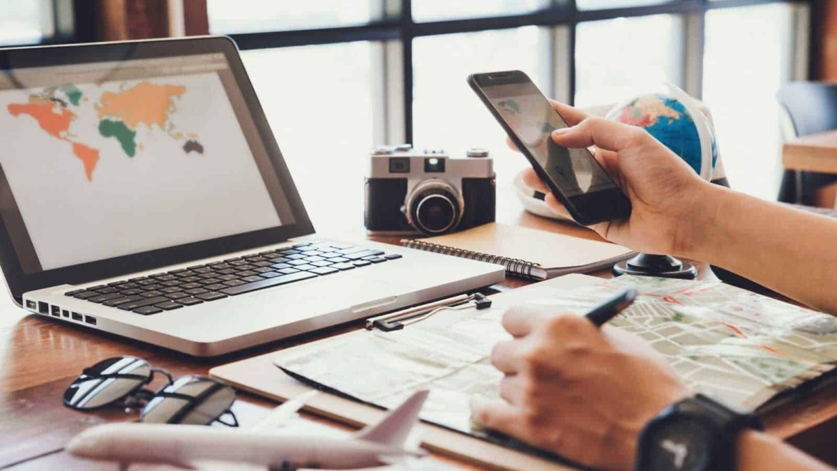 Viaggio e tecnologia: persona che utilizza smartphone e laptop in uno spazio di coworking con mappamondo, macchina fotografica e planning di viaggio.