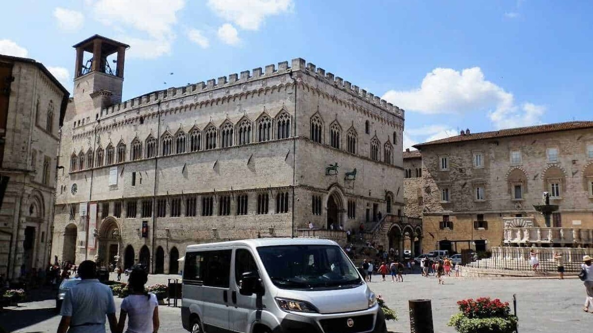 Antico palazzo medievale in un’affascinante piazza storica italiana con turisti, fiori e veicoli moderni, atmosfera autentica e architettura storica in una città affascinante.