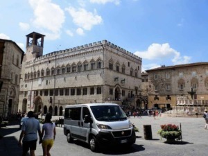 Antico palazzo medievale in un’affascinante piazza storica italiana con turisti, fiori e veicoli moderni, atmosfera autentica e architettura storica in una città affascinante.